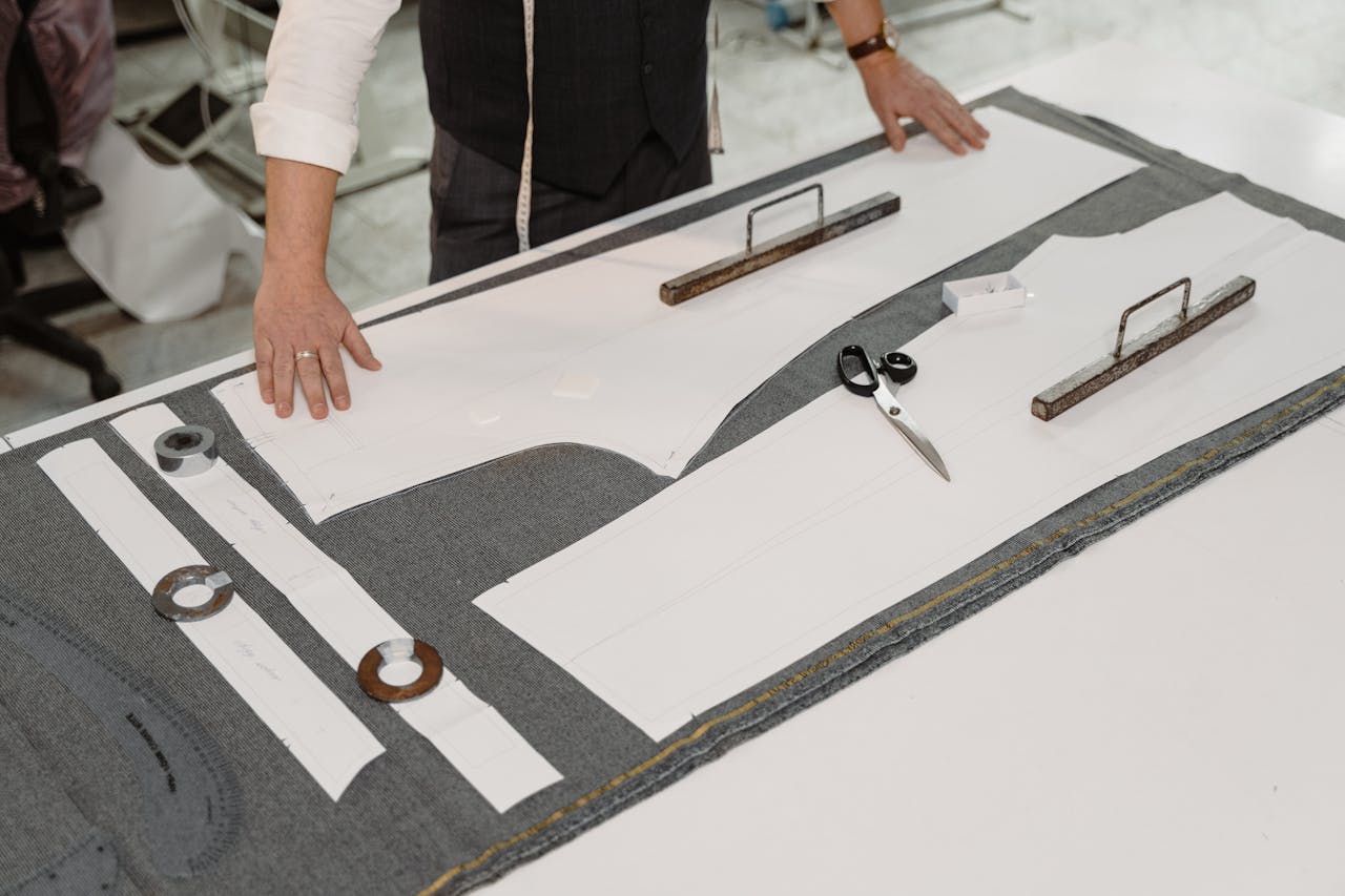 hero-img-02 Hands of a tailor working with fabric and patterns in a workshop, showcasing traditional craftsmanship.